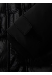 Primer plano de la manga de una chaqueta acolchada negra con una correa ajustable de Velcro sobre una tela acolchada brillante.