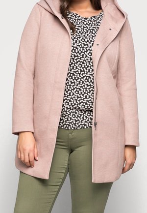Femme portant un manteau à capuche rose clair par-dessus un top floral noir et blanc et un pantalon vert olive, debout, les bras détendus le long du corps.