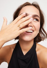Bague en or avec un design incurvé et une pierre précieuse centrale, portée sur une main avec des ongles naturels, sur un fond neutre.