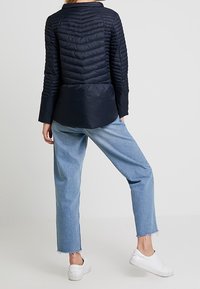 Chaqueta acolchada azul marino con un diseño estructurado, que cuenta con un cuello alto y una sección inferior lisa en contraste; combinada con unos jeans claros y holgados.