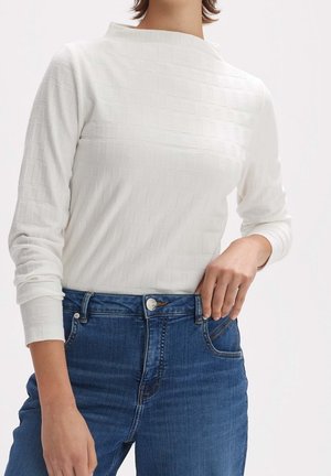 Kobieta w białej, teksturowanej bluzce z długim rękawem, włożonej do wysokich, niebieskich dżinsów, z ręką opartą blisko pasa.