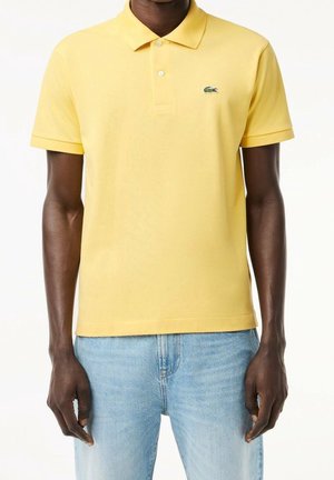 Polo jaune en coton, avec un col, trois boutons et un petit logo vert sur la poitrine gauche. Associé à un jean bleu clair.