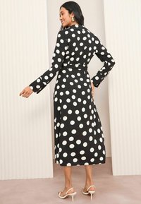 Schwarzes Kleid mit weißen Punkten, langen Ärmeln und tailliertem Bund. Hat eine Länge bis zur Mitte der Waden und ist aus einem glatten, leichten Stoff.