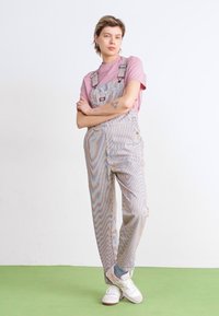 Un mono de rayas en colores claros, con tirantes ajustables, botones laterales y un corte relajado, combinado con una camiseta de manga corta rosa y zapatillas blancas.