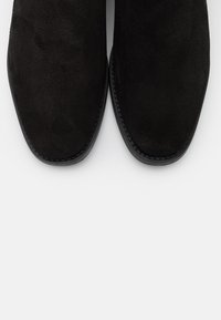 Bottes cheville en daim noir à finition lisse, bout carré, avec une couture contrastante le long de la semelle, posées sur un fond blanc.