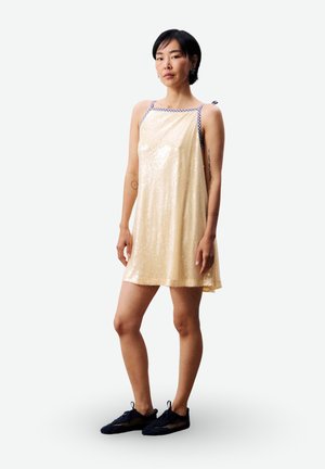 Femme debout en robe sans manches beige clair à paillettes avec bretelles vichy, portant des baskets noires, sur un fond blanc uni.