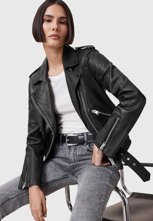 Veste de moto en cuir noir avec des zips en argent, des poches latérales et une coupe ajustée. Portée par-dessus une chemise blanche avec un jean gris clair.
