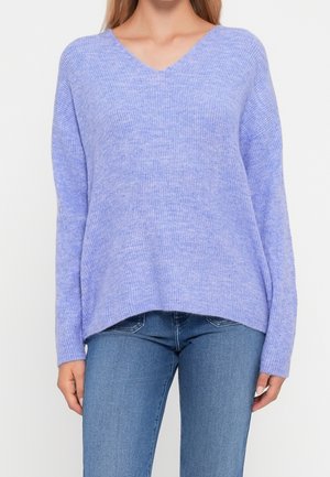 Kvinde iført en løs, ribstrikket lys lilla V-hals sweater kombineret med blå denimjeans, stående mod en ensfarvet baggrund.