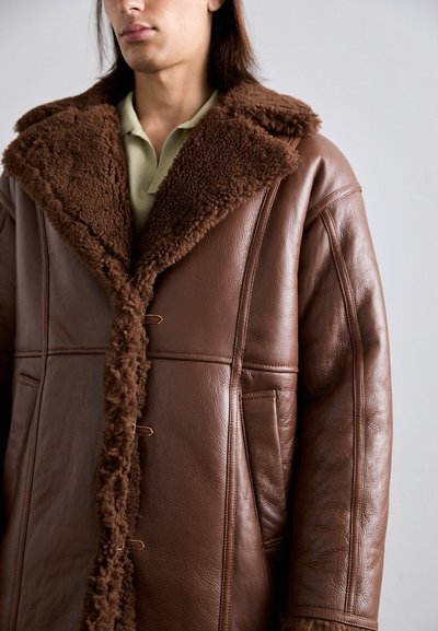 Persona che indossa un cappotto di pelle marrone con un ampio colletto in shearling testurizzato e rifiniture frontali, sopra una camicia col verde chiaro.