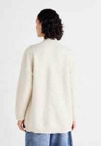 Krémoově bílý oversized svetr s texturovaným povrchem, žebrovanými manžetami a vysokým límcem. Design se vyznačuje uvolněným střihem a minimálním počtem švů.
