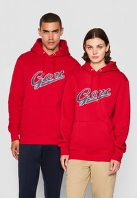 Røde hettegensere med en frontlomme og snor i hetten. Begge har "Gant"-logoet i marineblått over brystet.