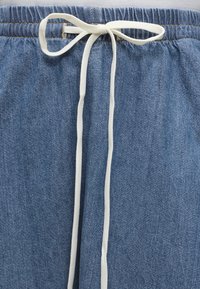 Μπλε τζιν παντελόνι με ελαστική μέση και λευκό κορδόνι δεμένο σε φιογκάκι στο κέντρο μπροστά.