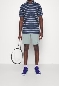 Polo rayé bleu marine à manches courtes, associé à un short vert clair. Tenant une raquette de tennis, portant des chaussures de sport violettes et vertes.