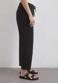 Zwarte, loszittende broek met een elastische tailleband en trekkoord. Draagt bij zwarte sandalen met gouden gespen en een gestructureerd oppervlak.