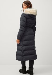 Manteau long noir en duvet avec une capuche bordée de fourrure, présentant des sections matelassées, une taille cintrée et une silhouette droite, associé à des bottes noires.