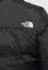 Veste matelassée noire avec logo "The North Face" brodé en blanc sur le haut du dos, près de l'épaule.