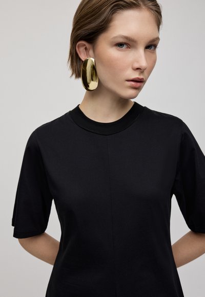 T-shirt nero a maniche corte con scollatura rotonda, caratterizzato da una texture liscia e una cucitura centrale. Una modella indossa grandi orecchini a cerchio dorati e lucenti.