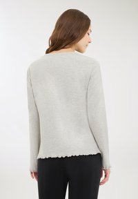 Una donna con lunghi capelli castani indossa un top grigio chiaro a maniche lunghe con bordi arricciati e pantaloni neri, voltata di spalle contro uno sfondo bianco.