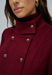Abrigo de lana burdeos con un alto cuello, diseño de doble botonadura y botones dorados. Presenta una textura suave y un corte ajustado.