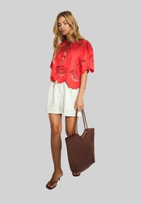 Crop top rosso ricamato con bordi a scallop, abbinato a pantaloni corti bianchi in lino e una borsa a tracolla intrecciata marrone. Il modello indossa ciabatte nere.