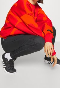 Sweatshirt rouge avec motif abstrait orange, leggings noirs à design texturé, et chaussures de sport noires avec des rayures blanches.