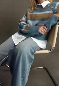 Pull rayé en laine mélangée bleu et marron avec des poignets côtelés et un détail de bouton doré. Pantalon large en jean bleu clair et chemise blanche en dessous.