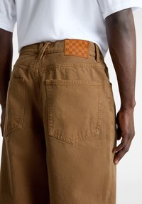 Hombre vestido con pantalones marrones con bolsillos traseros y una camisa blanca, visto de la cintura a la mitad del muslo, sobre un fondo liso.