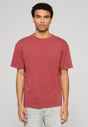 Jeune homme aux cheveux bouclés portant un t-shirt à manches courtes uni rouge rouille et un jean gris clair, debout devant un fond neutre.