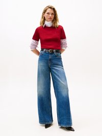 Suéter rojo de manga corta superpuesto a una camisa blanca con cuello, combinado con jeans azules de tiro alto y pierna ancha, y tacones negros de punta.