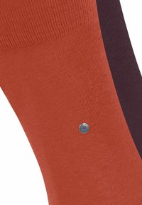 Zbliżenie skarpetki w pomarańczowych i ciemnofioletowych odcieniach, z metalowym emblematem po boku, podkreślające teksturę i kontrast kolorów.