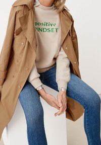 Beige trenchcoat över ljus sweatshirt med "positiv MINDSET" i grönt, kombinerat med denimjeans. Sweatshirten har blommiga detaljer på ärmarna.