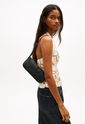 Γυναίκα που φοράει τοπ με λουράκι στον λαιμό και σχέδιο με αλυσίδες, καθώς και σκούρο τζιν, κρατώντας μια μαύρη τσάντα ώμου Tommy Jeans.