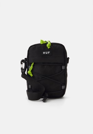 Geantă crossbody din material textil negru, cu accente din sfoară verde, un patch cu logo și bretele ajustabile. Prezintă o suprafață texturată și cordoane elastice.