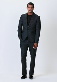 Schwarzer Anzug mit geometrischem Muster, bestehend aus einem einreihigen Blazer und maßgeschneiderten Hosen. Glänzende schwarze Schuhe runden das Outfit ab.