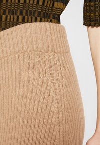 Gros plan de la ceinture d'une jupe en tricot côtelé beige associée à un haut marron et noir à motifs, avec un bras visible sur le côté droit.