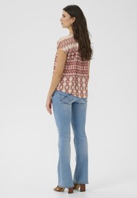 Bluse mit floralem Muster in Rost und Creme mit kurzen Ärmeln, kombiniert mit hellblauen, eng sitzenden Jeans. Die Bluse hat einen abgerundeten Saum und eine lockere Passform.
