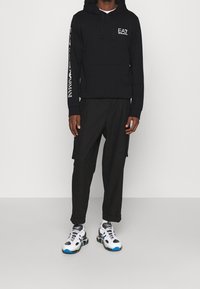 Sudadera negra con bolsillo frontal y logo, acompañada de pantalones cargo negros. Los zapatos tienen un diseño en blanco y negro con acentos azules.