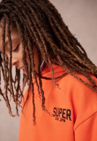 Person med lange dreadlocks iført en orange sweatshirt med "SUPER DRY JPN" broderet på brystet, vendt nedad.