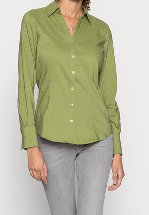 Kobieta ubrana w dopasowaną oliwkowozieloną koszulę na długi rękaw z kołnierzykiem oraz jasnoszare dżinsy, stojąca na jednolitym tle.