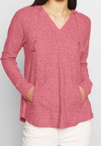 Hooded longsleeve top in gemêleerd roze stof. Bevat een V-hals, franjes aan de kap en voorzakken. Zachte textuur, ontspannen pasvorm.