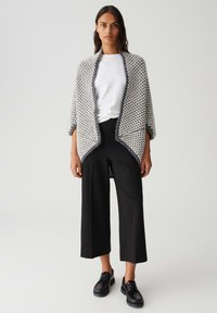 Cardigan tricoté gris et blanc à motif avec des bords frangés, porté sur une chemise blanche et un pantalon noir à jambes larges, associé à des chaussures noires.