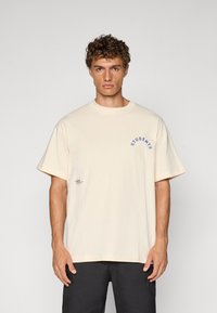 Beige bomulls t-skjorte med løs passform, rund hals, og "STUDENTS" i blå båtrykk på venstre bryst. Inkluderer en liten etikett på siden.