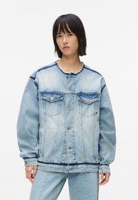 Cilvēks, kas valkā pārāk lielu gaiši zilu denim jaku un saskaņot�ās bikses, stāvot pretī vienkāršai baltai fona.