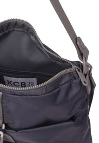 Bolsa de tela de color gris oscuro con apertura con cremallera, textura suave y correa ajustable. Bolsillo interior visible con etiqueta de la marca.