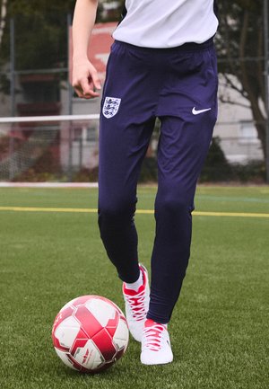 Pantalons de sport bleu marine avec un emblème d'Angleterre et un logo Nike blanc ; portés avec des chaussures de football blanches et rouges ; joueur donnant un coup de pied à un ballon de football rouge et blanc.