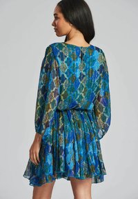 Vestido de manga corta en tejido estampado de color azul, verde y dorado. Presenta una textura plisada y una abertura en forma de llave en la parte posterior. Línea de dobladillo acampanada.