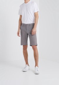 Uomo che indossa una maglietta bianca semplice, pantaloni corti grigi fino al ginocchio e sneakers bianche, in piedi contro uno sfondo bianco.