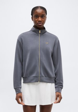 Kobieta ubrana w szarą kurtkę zapinaną na suwak z złotym suwakiem i logo EA7, w połączeniu z białą spódnicą, stojąca na jednolitym tle.