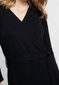 Musta kietaisumekko, jossa on V-pääntie, pitkät hihat ja vyötäröllä solmittava vyö, valmistettu pehmeästä ja sileästä kankaasta.