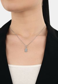 Collana d'argento con pendente rettangolare caratterizzato da piccole linee incrociate, indossata al collo di una donna sopra una maglia bianca e una giacca nera.